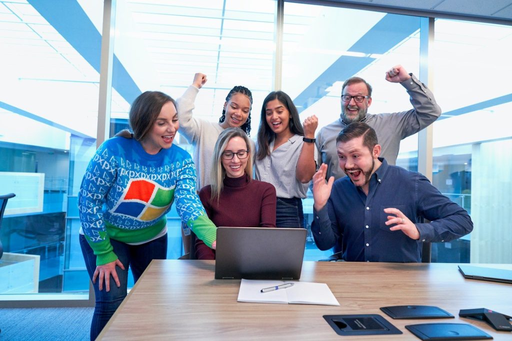 Your Success Story: How To Be A Winner In Business 2 men and women sitting and standing by the table looking happy while staring at laptop